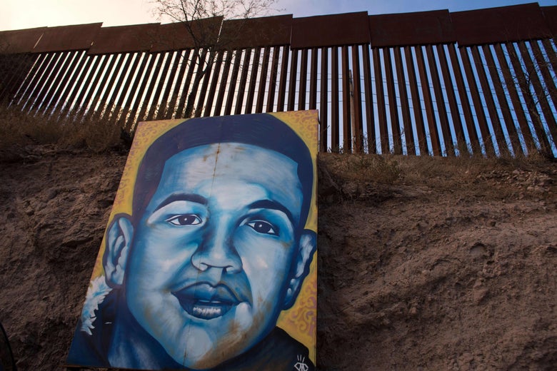A large portrait of a teenage boy, painted in blues on a yellow background, leans against earth under a border fence.